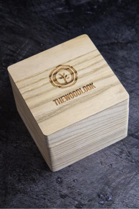 A square light wood box with a laser-engraved logo and the brand name 'THEWOODLOOK' on the top, set against a dark textured background.