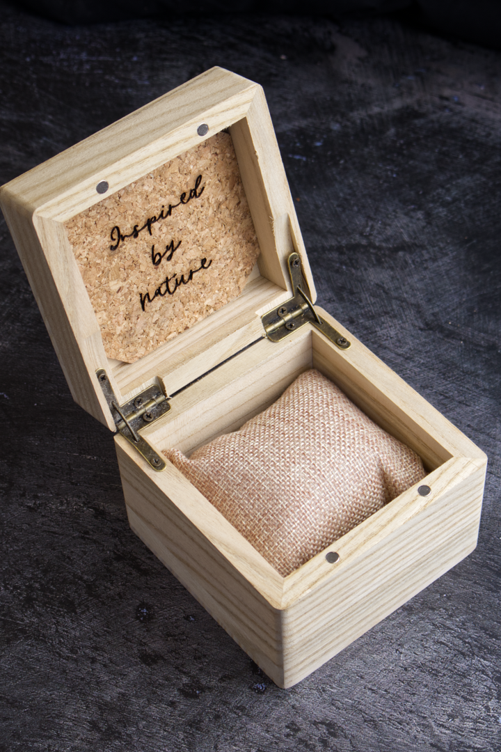 A small open wooden keepsake box with a burlap-lined interior and a cork insert on the lid that reads 'Spend it on nature.' The box sits on a dark textured surface.